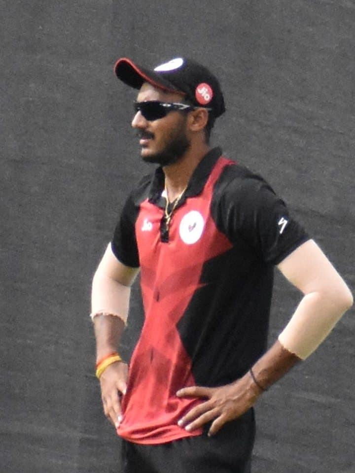Axar Patel in Gujarat's red and black kit and sunglasses, hands on hips, fielding on the boundary.