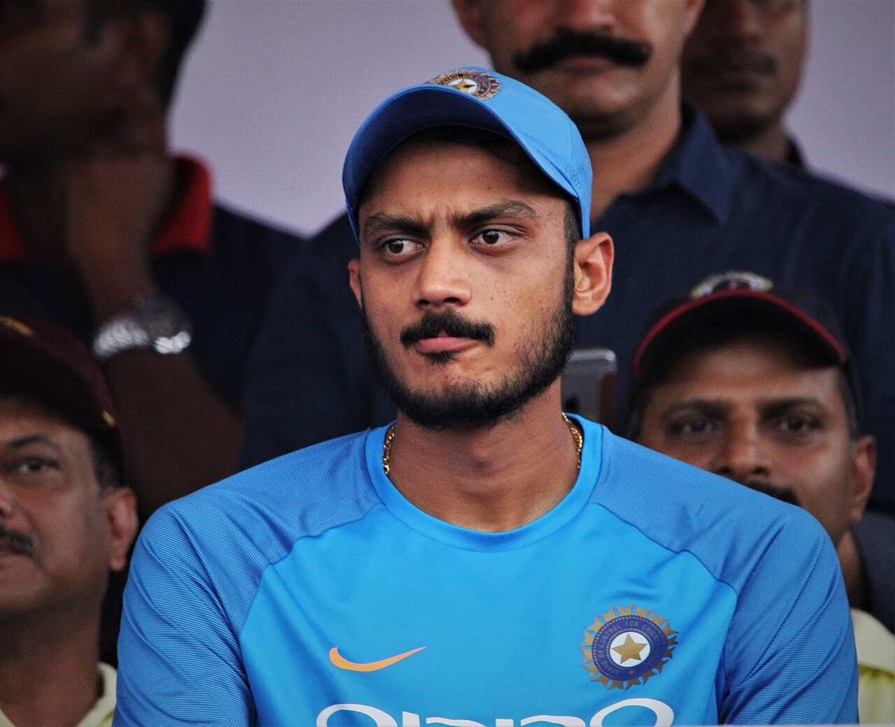 Axar Patel in an India training shirt and blue cap, seated among spectators in a stand.